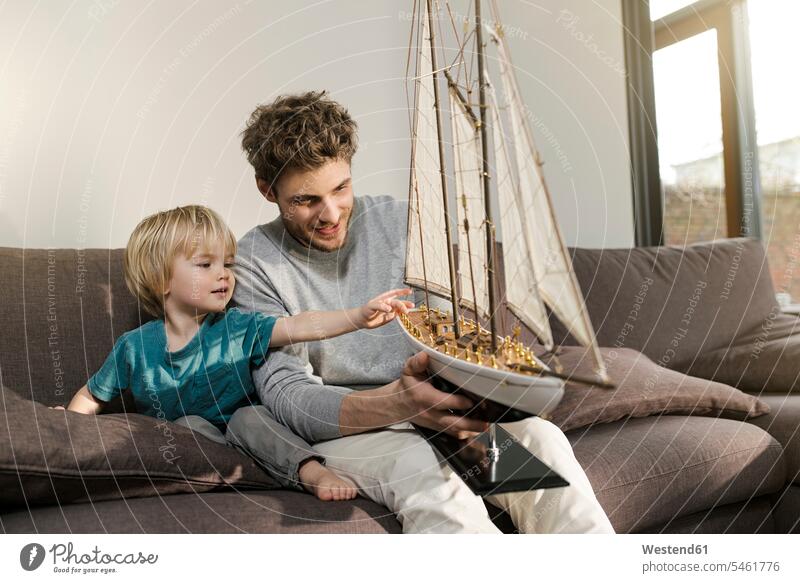 Vater und Sohn schauen sich zu Hause auf der Couch ein Spielzeug-Modellschiff an Zuhause daheim Söhne Sofa Couches Liege Sofas sitzen sitzend sitzt Modellboot