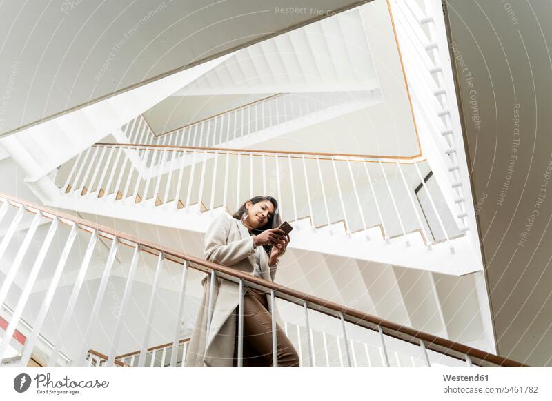 Junge Frau mit Smartphone geht im Treppenhaus die Treppe hinunter Informationen Telekommunikation telefonieren Handies Handys Mobiltelefon Mobiltelefone gehend
