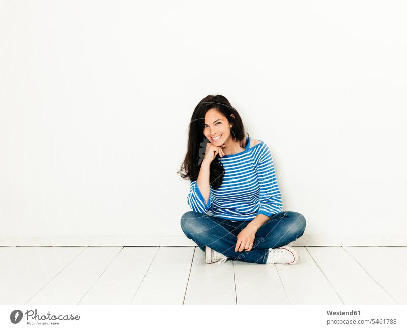 Schöne junge Frau mit schwarzen Haaren und blau-weiß gestreiften Pullover sitzt auf dem Boden vor dem weißen Hintergrund Europäer Kaukasier Europäisch