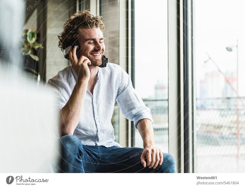 Glücklicher lässiger junger Mann, der mit Kopfhörern am Fenster Musik hört glücklich glücklich sein glücklichsein leger Männer männlich hören hoeren Kopfhoerer