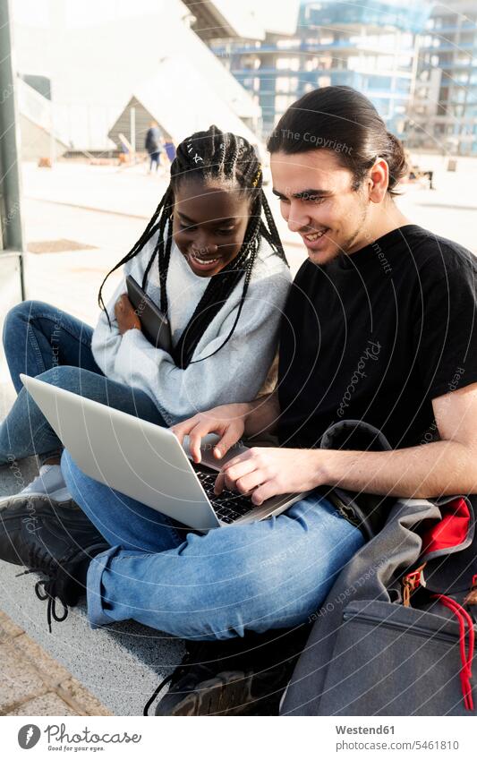 Glückliches junges Paar mit Laptop an der Straßenbahnhaltestelle Freunde Kameradschaft Rucksäcke Hosen Jeanshose Bänke Sitzbank Sitzbänke Rechner Laptops