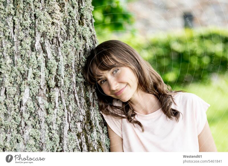 Porträt einer lächelnden brünetten Frau, die sich gegen einen Baumstamm lehnt Leute Menschen People Person Personen Europäisch Kaukasier kaukasisch 1 Ein