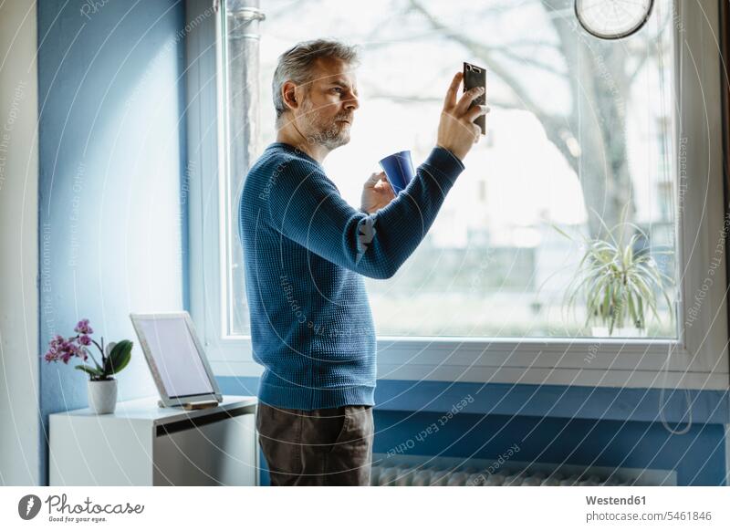 Älterer Mann mit Kaffeetasse im Wohnzimmer stehend, der ein Selfie mit seinem Smartphone macht Leute Menschen People Person Personen Europäisch Kaukasier