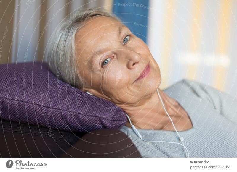 Porträt einer lächelnden älteren Frau, die auf der Couch zu Hause liegt und mit Kopfhörern Musik hört weiblich Frauen Seniorin Seniorinnen alt Zuhause daheim
