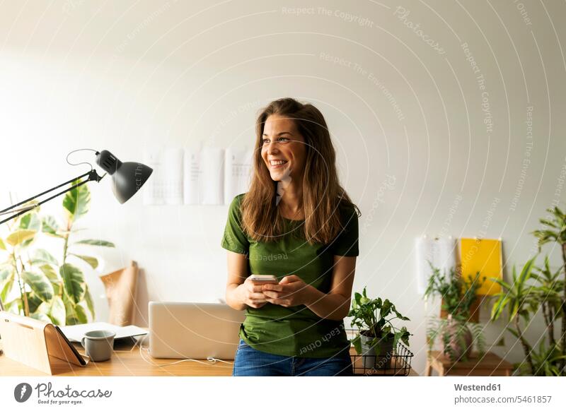 Lächelnde junge Frau hält Smartphone in der Hand und schaut weg, während sie am Schreibtisch im Home-Office steht Farbaufnahme Farbe Farbfoto Farbphoto Spanien