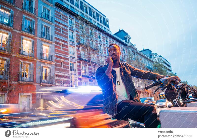 Lächelnder Mann spricht über Smartphone, während er in der Dämmerung in der Stadt auf dem Fahrrad sitzt Farbaufnahme Farbe Farbfoto Farbphoto Spanien