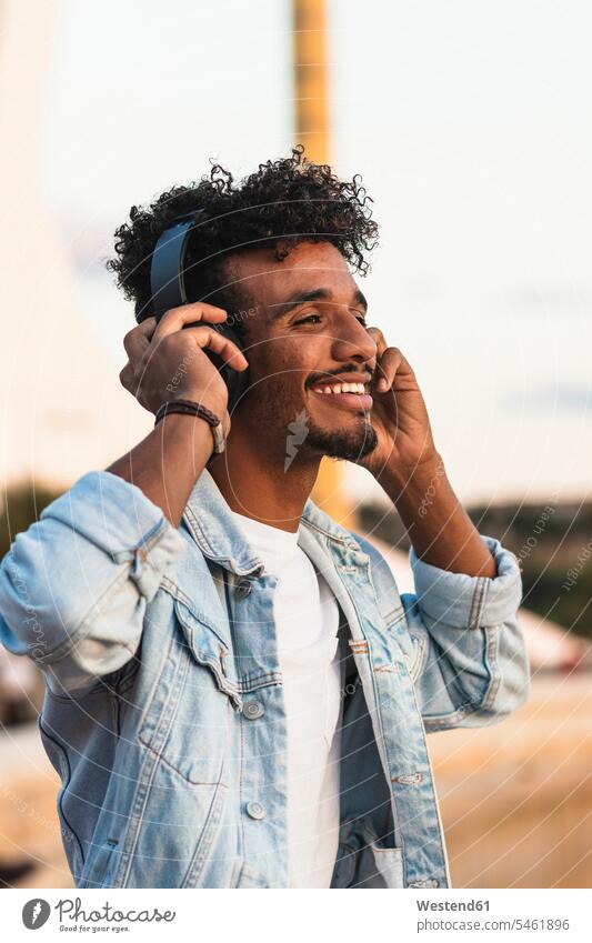 Nahaufnahme eines lächelnden jungen Mannes, der über Kopfhörer Musik gegen den klaren Himmel hört Farbaufnahme Farbe Farbfoto Farbphoto Spanien