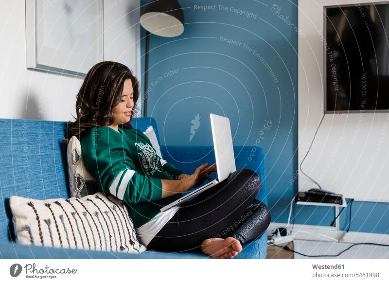 Junge Frau mit Down-Syndrom, die einen Laptop benutzt, während sie zu Hause auf dem Sofa im Wohnzimmer sitzt Farbaufnahme Farbe Farbfoto Farbphoto Innenaufnahme