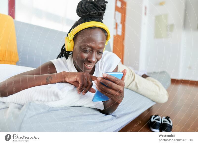Junge Frau, die mit einem Smartphone über Kopfhörer Musik hört, während sie zu Hause auf dem Sofa liegt Farbaufnahme Farbe Farbfoto Farbphoto Freizeitkleidung