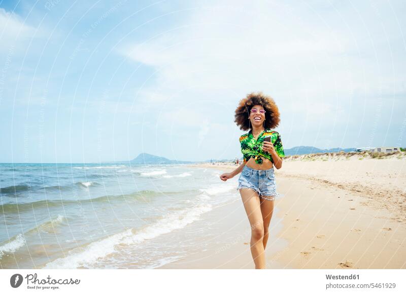 Junge Frau rennt am Strand Telekommunikation telefonieren Handies Handys Mobiltelefon Mobiltelefone Brillen Sonnenbrillen rennen Glück glücklich sein