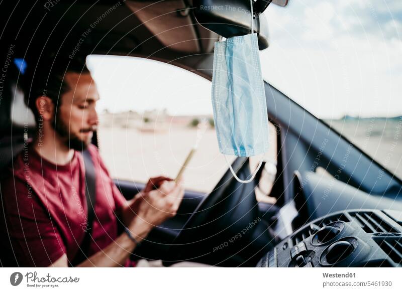Nahaufnahme einer Gesichtsmaske, die am Rückspiegel hängt, während ein Mann im Auto telefoniert Farbaufnahme Farbe Farbfoto Farbphoto Fahrzeuginnenraum Tag