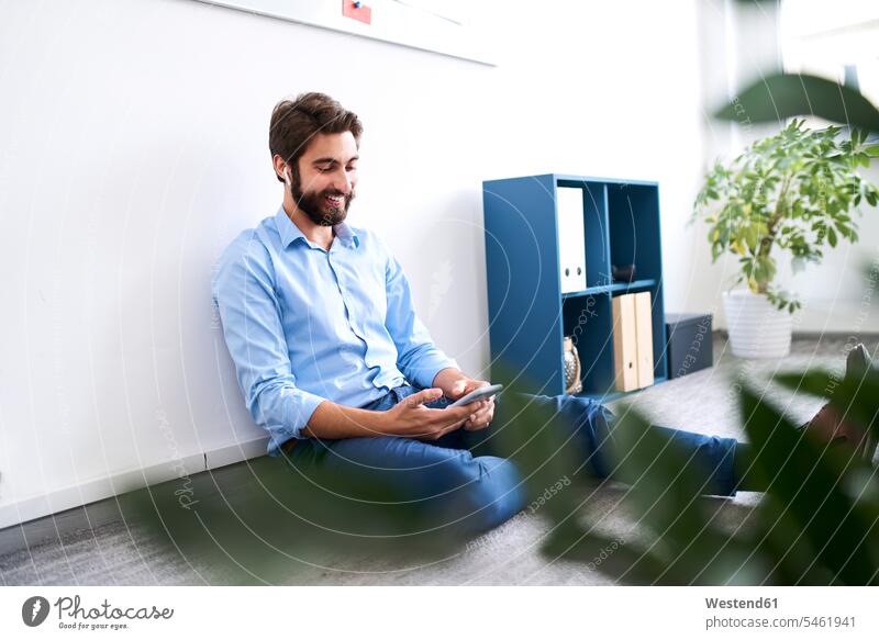 Lächelnder Geschäftsmann, der auf dem Boden sitzt und sein Smartphone benutzt Job Berufe Berufstätigkeit Beschäftigung Jobs Designer geschäftlich Geschäftsleben