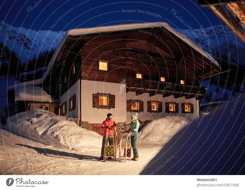 Paar mit Schlitten in schneebedeckter Landschaft mit rustikalem Haus bei Nacht Landschaften Häuser Haeuser nachts Pärchen Paare Partnerschaft verschneit Gebäude