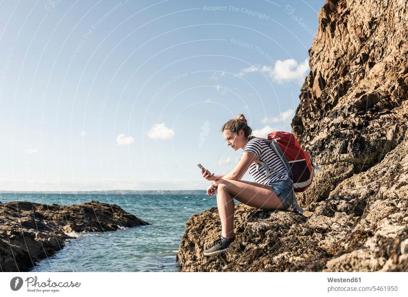 Junge Frau sitzt auf einem felsigen Strand, mit Smartphone Strandwanderung Fels Felsen sitzen sitzend benutzen benützen Beach Straende Strände Beaches iPhone