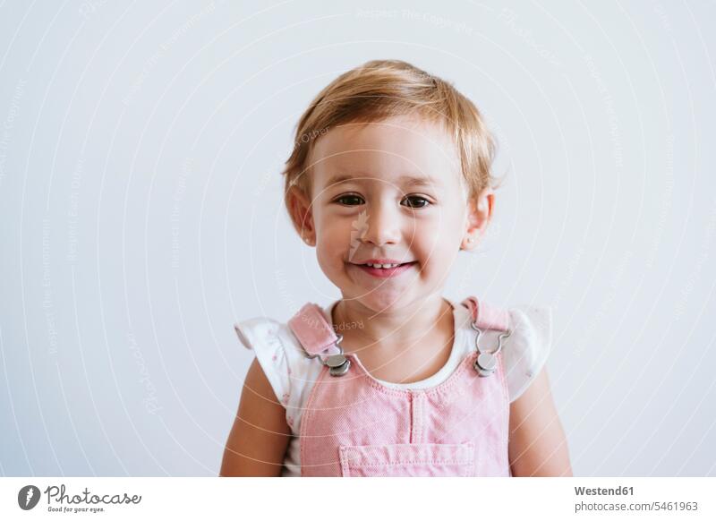 Lächelndes kleines Mädchen steht an weißer Wand Farbaufnahme Farbe Farbfoto Farbphoto Innenaufnahme Innenaufnahmen innen drinnen Tag Tageslichtaufnahme