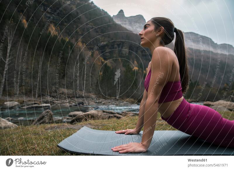 Aktive junge Frau übt Kobra-Pose auf Yogamatte in der Nähe von Fluss und Berg Farbaufnahme Farbe Farbfoto Farbphoto Außenaufnahme außen draußen im Freien Huesca