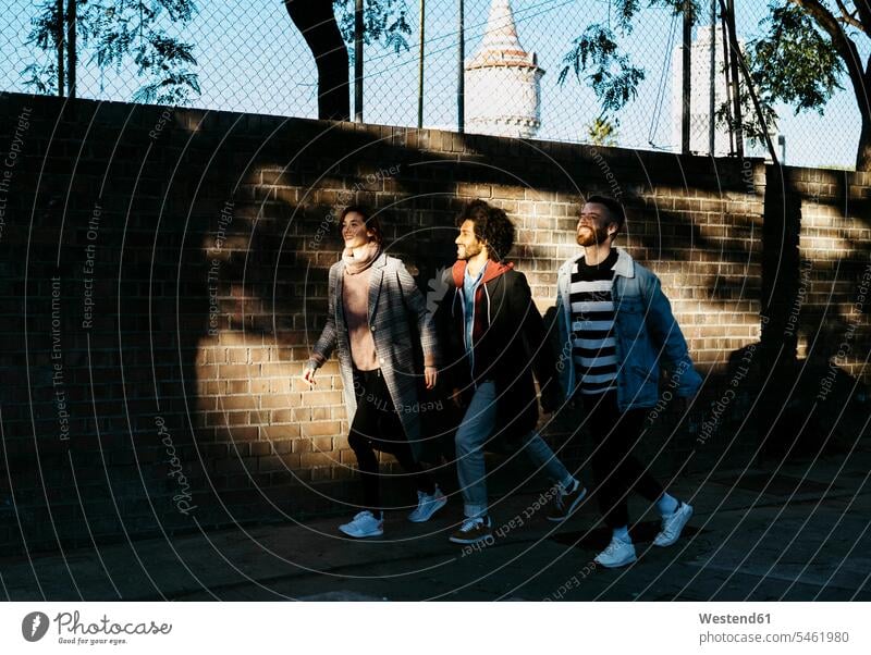 Drei glückliche Freunde gehen entlang einer Ziegelmauer im Schatten Glück glücklich sein glücklichsein gehend geht Backsteinwand Backsteinmauern Freundschaft