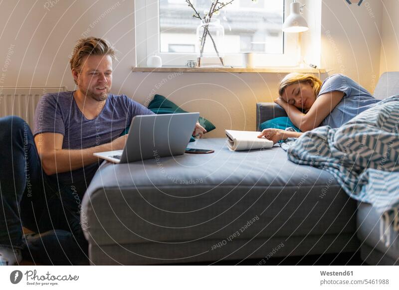 Junge Frau, die zu Hause mit ihrem Partner auf der Couch liegt und einen Laptop benutzt Decken Beleuchtungen Lampen Leuchte Bücher Couches Liege Sofas Rechner