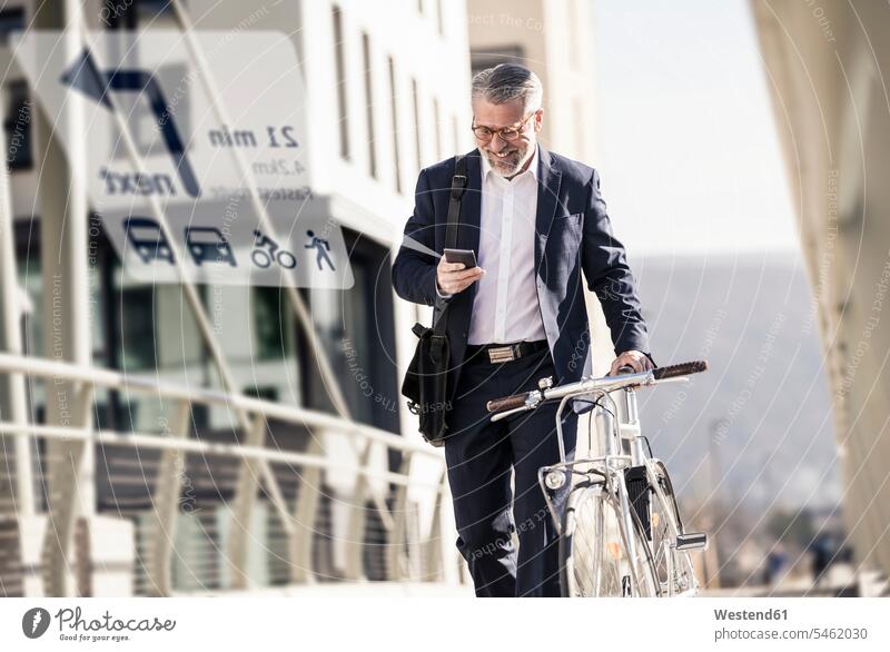 Lächelnder reifer Geschäftsmann mit Fahrrad, der sein Handy zur Navigation in der Stadt benutzt Leute Menschen People Person Personen Europäisch Kaukasier