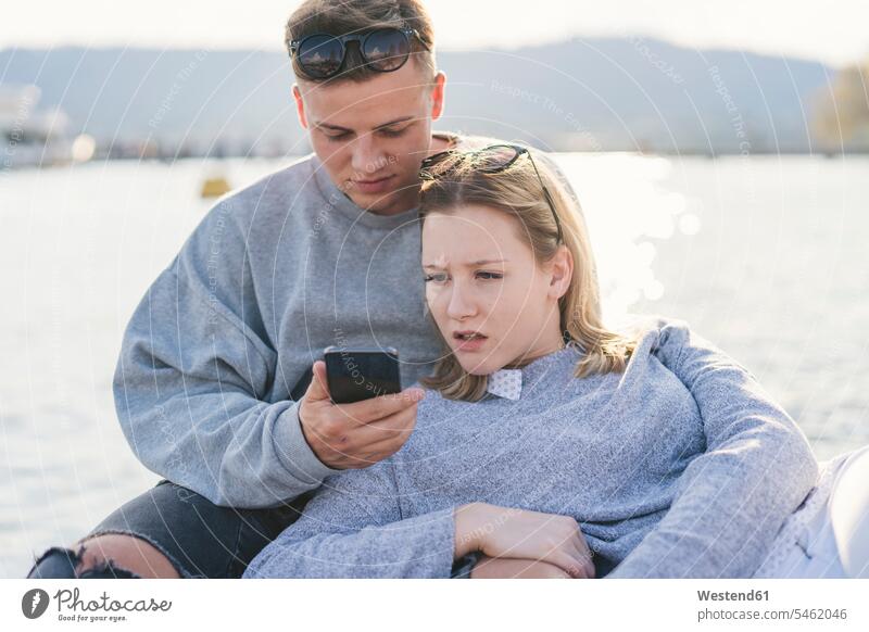 Junges Paar auf Steg am Zürichsee schaut auf Handy, Zürich, Schweiz Informationen Telekommunikation Handies Handys Mobiltelefon Mobiltelefone entdecken Lektüre