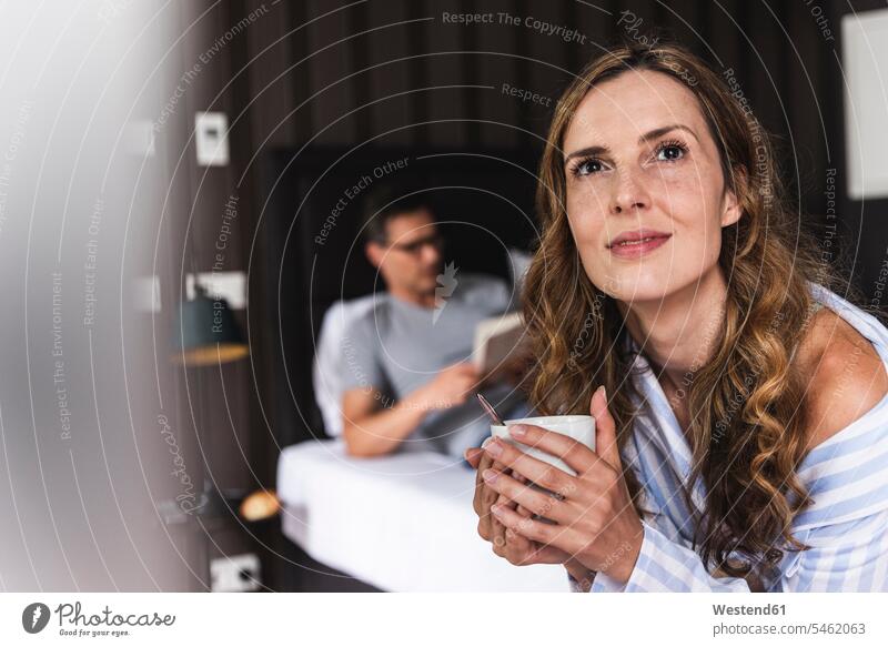 Lächelnde Frau mit einer Tasse Kaffee im Schlafzimmer mit einem Mann im Hintergrund Zuhause zu Hause daheim lächeln weiblich Frauen Zimmer Raum Räume Getränk