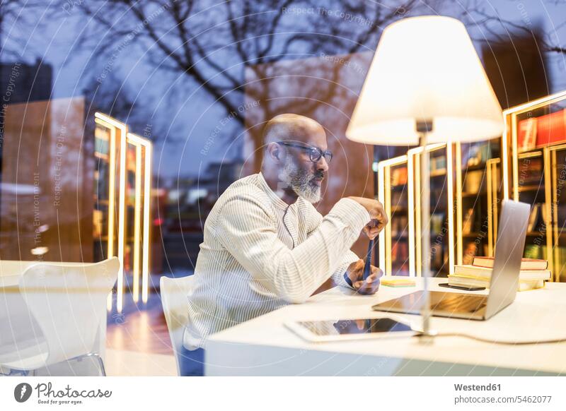 Älterer Mann sitzt am Schreibtisch in einer Bibliothek am Abend mit Laptop Notebook Laptops Notebooks abends benutzen benützen Arbeitstisch Schreibtische Männer