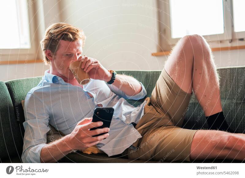 Mann trinkt Kaffee, während er ein Smartphone im Wohnzimmer benutzt Farbaufnahme Farbe Farbfoto Farbphoto Innenaufnahme Innenaufnahmen innen drinnen Tag