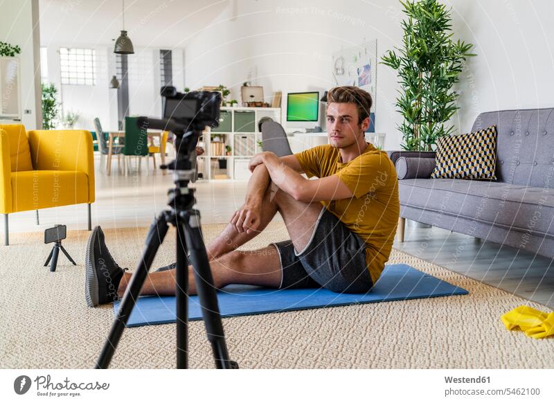 Fitnesstrainer, der zu Hause auf der Matte sitzend die Übung mit einer Kamera aufzeichnet Farbaufnahme Farbe Farbfoto Farbphoto Innenaufnahme Innenaufnahmen