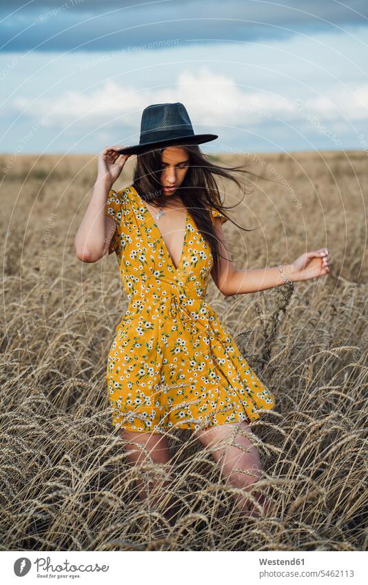 Junge Frau trägt ein Sommerkleid mit Blumenmuster und einen Hut, der im Maisfeld tanzt Hüte Kornfeld Getreidefeld Getreidefelder Kornfelder Feld Felder