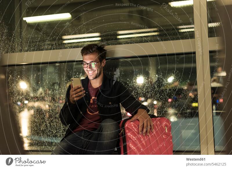 Lächelnder junger Mann mit Koffer sitzt am verregneten Fenster und benutzt ein Smartphone Leute Menschen People Person Personen Europäisch Kaukasier kaukasisch