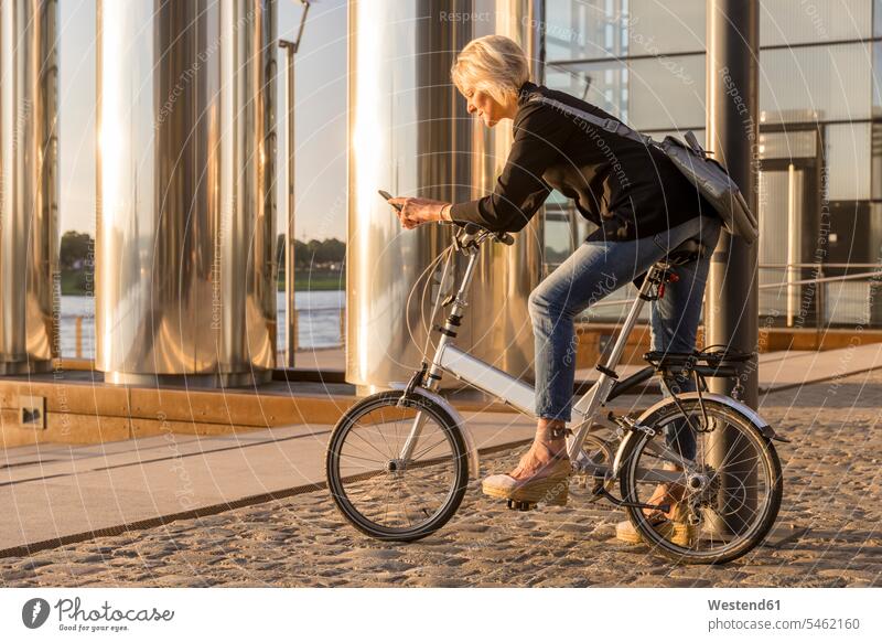 Ältere Frau mit Stadtfahrrad mit Handy am Flussufer bei Sonnenuntergang Fahrrad Bikes Fahrräder Räder Rad weiblich Frauen Sonnenuntergänge Mobiltelefon Handies