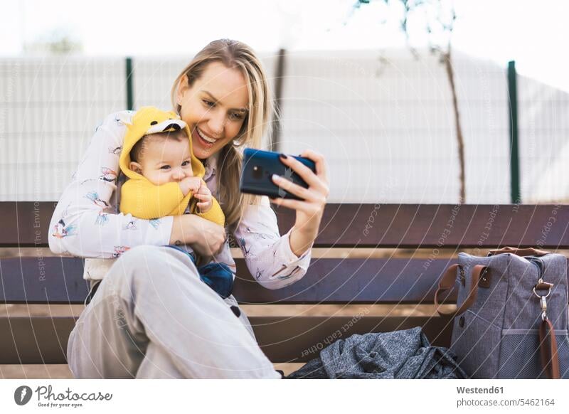 Glückliche Mutter ruht mit Baby auf einer Parkbank und macht ein Selfie Taschen Jacken Bänke Sitzbank Sitzbänke Parkbänke Telekommunikation telefonieren Handies