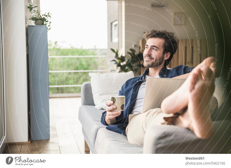Junger Mann sitzt auf Couch, benutzt Laptop, trinkt Kaffee Deutschland Laptop benutzen Laptop benützen Drahtlose Technologie drahtlose Verbindung
