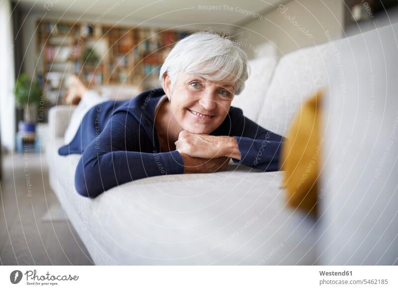 Lächelnde ältere Frau mit Hand am Kinn, die zu Hause auf dem Sofa liegt Farbaufnahme Farbe Farbfoto Farbphoto Innenaufnahme Innenaufnahmen innen drinnen Tag