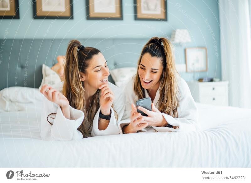 Lächelnde Zwillingsschwestern benutzen ein Smartphone, während sie im Hotel auf dem Bett liegen Farbaufnahme Farbe Farbfoto Farbphoto Spanien
