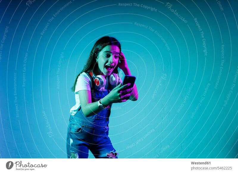 Aufgeregtes Mädchen schaut auf Smartphone, während sie vor blauem Hintergrund steht Farbaufnahme Farbe Farbfoto Farbphoto Studioaufnahme Studioaufnahmen
