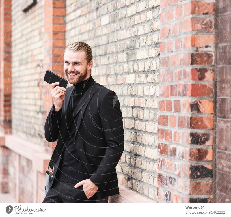 Lächelnde modischen jungen Mann steht an Backsteinmauer mit Handy lächeln Backsteinwand Backsteinmauern Portrait Porträts Portraits Mobiltelefon Handies Handys