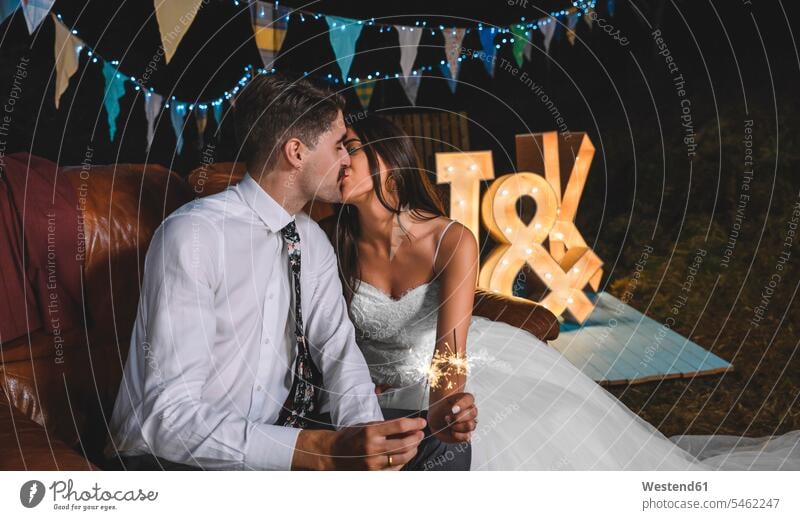 Romantisches Hochzeitspaar sitzt auf dem Sofa und küsst sich, während es auf einer nächtlichen Party im Freien Wunderkerzen in den Händen hält sitzen sitzend