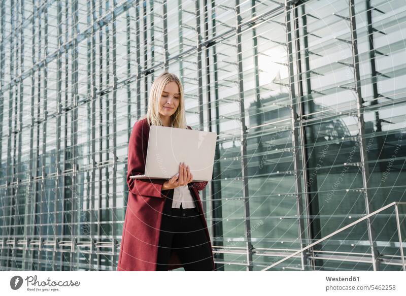 Blonde Geschäftsfrau mit Laptop im Hintergrund eines modernen Bürogebäudes Job Berufe Berufstätigkeit Beschäftigung Jobs geschäftlich Geschäftsleben