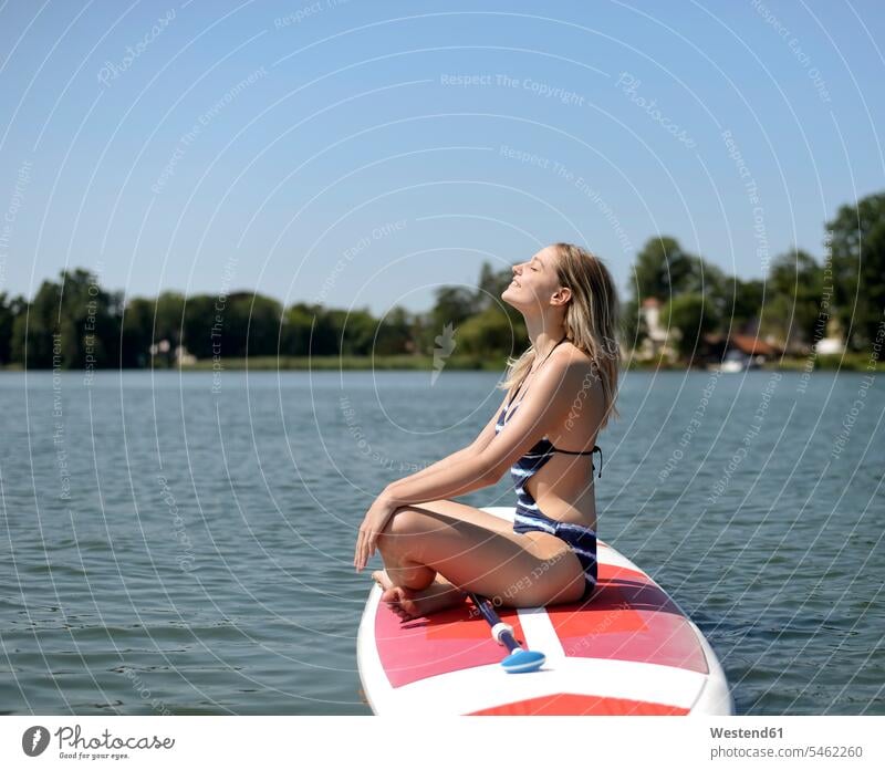 Deutschland, Brandenburg, Frau entspannt auf Paddelbrett auf Zeuthener See sitzen sitzend sitzt Seen lächeln Paddleboard weiblich Frauen blond blonde Haare
