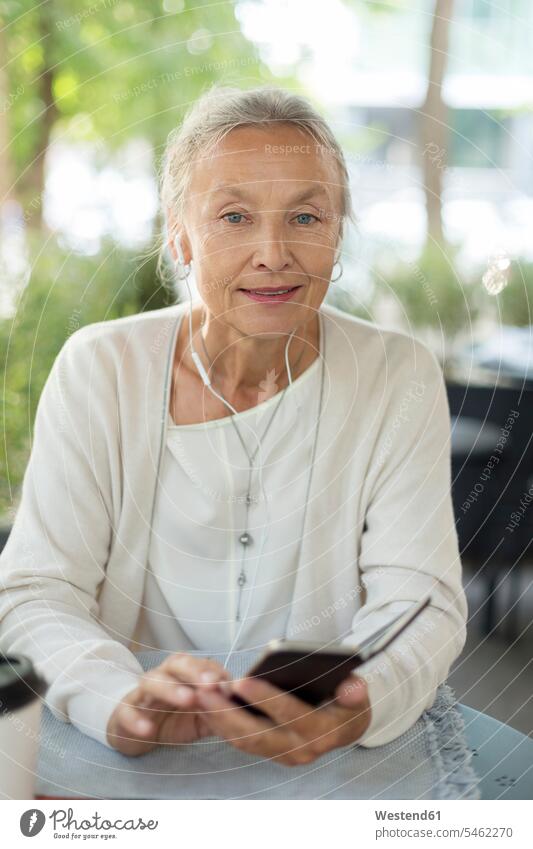 Porträt einer älteren Frau in einem Café im Freien mit Mobiltelefon und Kopfhörern Seniorin Seniorinnen alt Ohrhörer weiblich Frauen lächeln Handy Handies