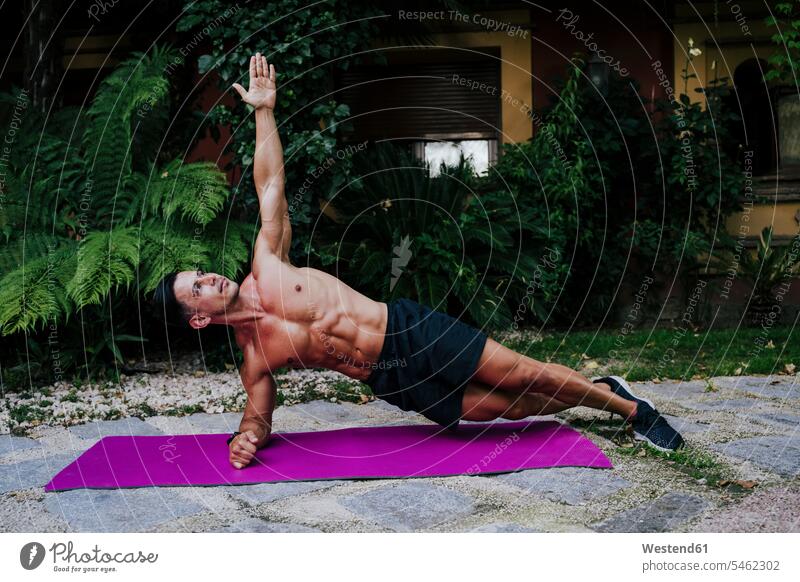 Männlicher Athlet ohne Hemd, der auf der Matte gegen Pflanzen im Hof die Plankenposition übt Farbaufnahme Farbe Farbfoto Farbphoto Spanien Außenaufnahme außen