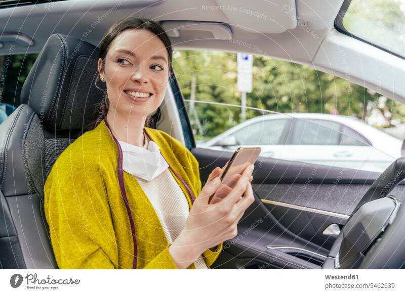 Porträt einer lächelnden Frau im Auto mit Schutzmaske und Smartphone Jacken KFZ Verkehrsmittel Automobil Autos PKW PKWs Wagen Telekommunikation Handies Handys