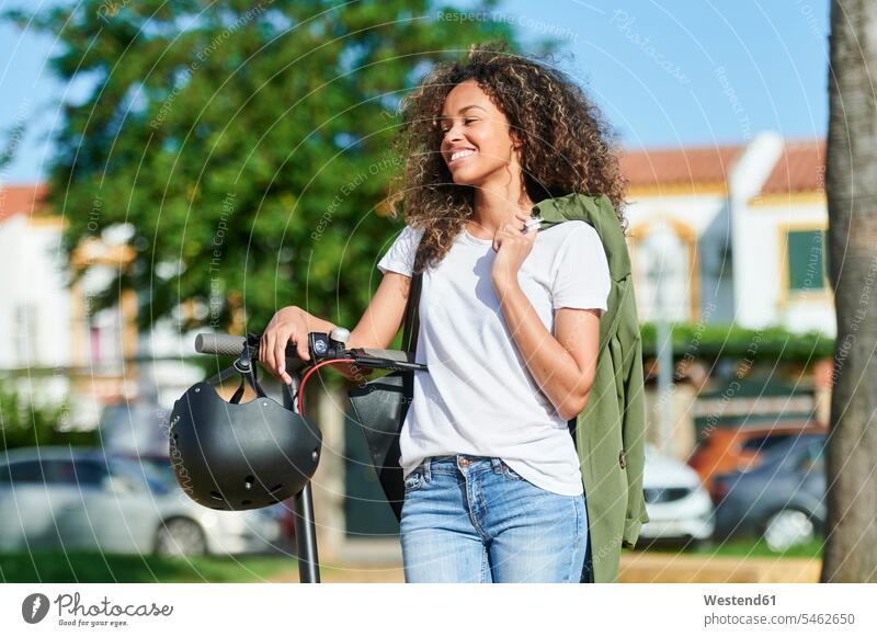 Glückliche Frau trägt Jacke, während sie an einem sonnigen Tag mit einem elektrischen Roller steht Farbaufnahme Farbe Farbfoto Farbphoto Außenaufnahme außen