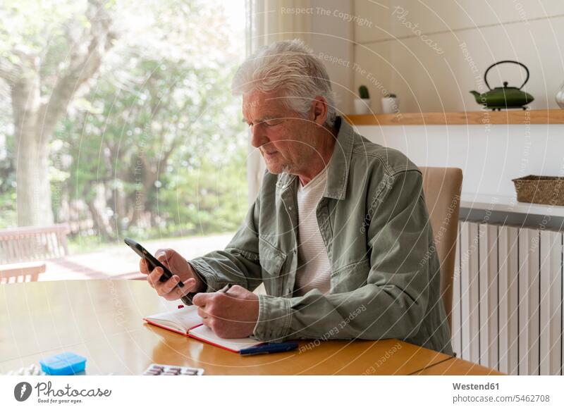 Älterer diabetischer Mann benutzt Mobiltelefon, während er zu Hause in sein Tagebuch schreibt Farbaufnahme Farbe Farbfoto Farbphoto Innenaufnahme Innenaufnahmen