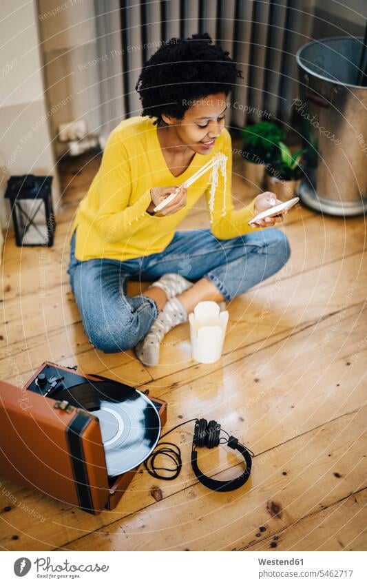 Junge Frau zu Hause, die mit einem Plattenspieler sitzt und während des Nudelessens das Smartphone kontrolliert Zuhause daheim junge Frau junge Frauen essend