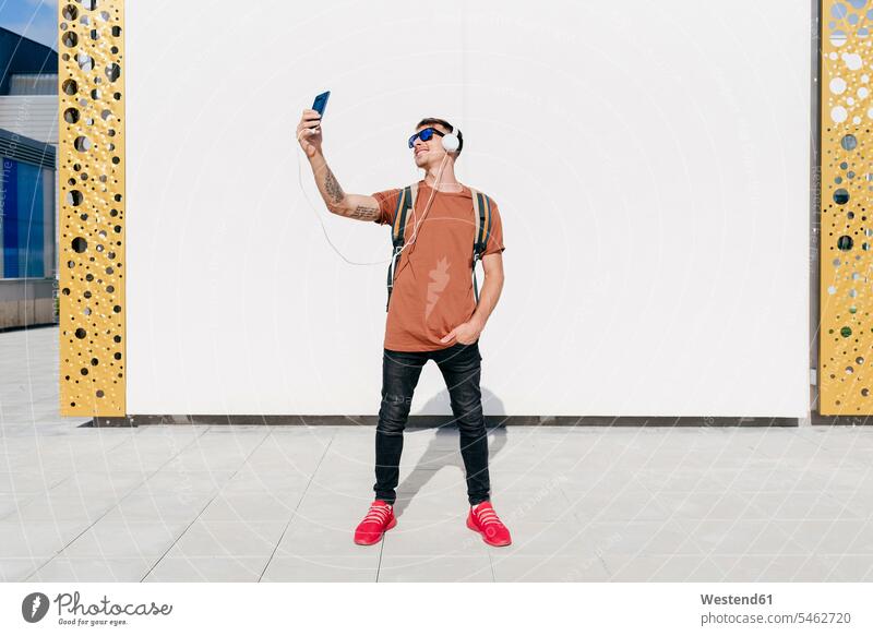 Lächelnder Mann mit Kopfhörern, der ein Selfie mit seinem Mobiltelefon macht, während er mit den Händen in den Taschen an der Wand steht Farbaufnahme Farbe