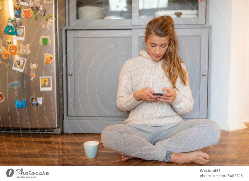 Junge Frau sitzt zu Hause in der Küche auf dem Boden und benutzt ihr Handy Leute Menschen People Person Personen Alleinstehende Alleinstehender Singles