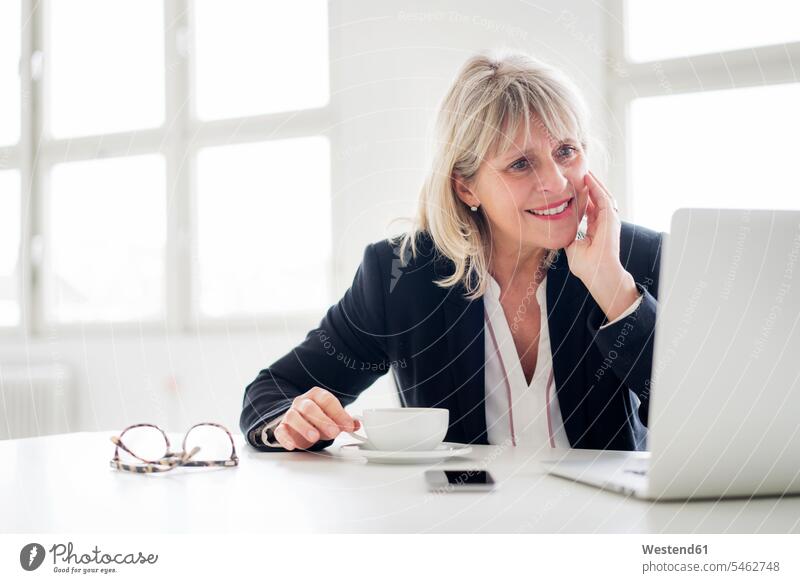 Lächelnde reife Geschäftsfrau mit einer Tasse Kaffee, die am Laptop am Schreibtisch im Büro arbeitet Notebook Laptops Notebooks Kaffeetasse Kaffeetassen