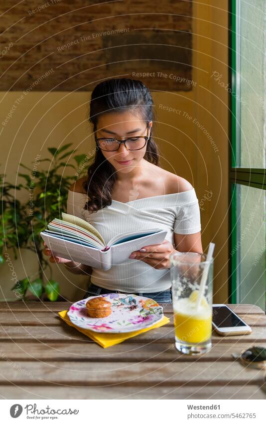 Junge Frau sitzt am Tisch und liest ein Buch Tische Holztische Lektüre Essen Essen und Trinken Food Lebensmittel Nahrungsmittel Leute Menschen People Person